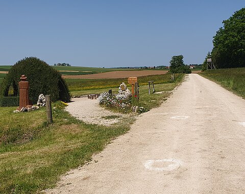 Ländlicher Weg mit Skulptur und Blumen, umgeben von Feldern und Bäumen unter klarem Himmel.
