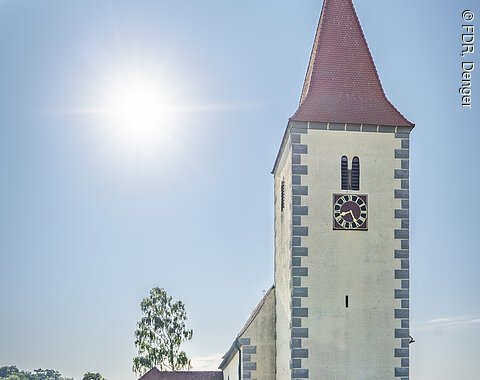 Kirchturm mit rotem Dach und Uhr, umgeben von einem Friedhof, unter klarem Himmel mit Sonne.