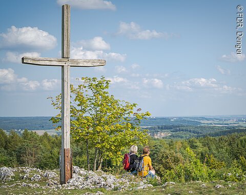 Aussicht Bockberg bei Harburg Holzkreuz auf dem Harburger Bockberg, zwei Personen mit Rucksäcken sitzen daneben, Blick auf die bewaldete Landschaft.