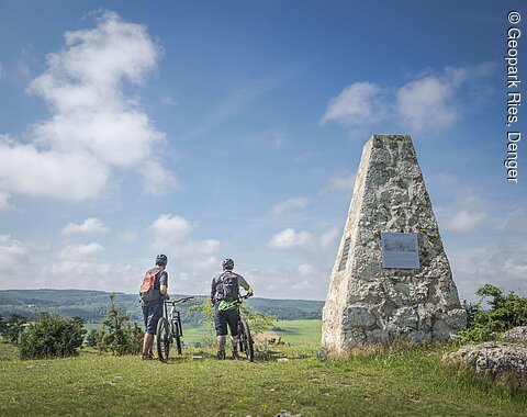 Denkmal auf dem Albuch am Geopark-Schwedenweg Zwei Radfahrer mit Helmen stehen neben einem steinernen Denkmal auf dem Albuch bei Hürnheim und blicken in die Ferne.