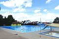 Freibad Monheim Ein Freibad mit einem Schwimmbecken, einer Rutsche und einem Kinderbecken. Im Hintergrund steht ein grünes Gebäude mit rotem Dach. Dahinter sind Wohnhäuser und Bäume und blauer Himmel zu sehen.