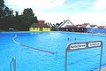 Freibad Monheim - Schwimmbecken für Nichtschwimmer und Schwimmer Ein Freibad mit einem großen Schwimmbecken, unterteilt in Nichtschwimmer- und Schwimmerbereich. Im Hintergrund sind Gebäude und Bäume zu sehen.
