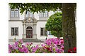 Eingang des Residenzschlosses mit verzierten Türen, umgeben von blühenden Blumen und Bäumen.