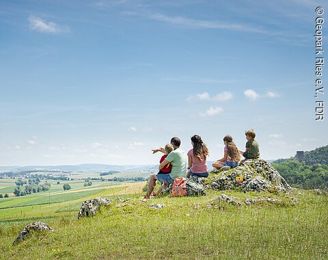Eine Gruppe von Menschen sitzt auf einem Hügel und blickt auf eine weite, grüne Landschaft.