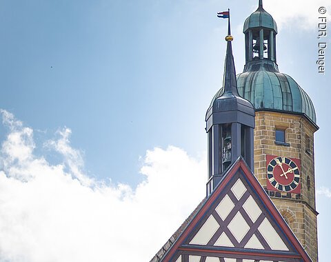 Auf dem Bild ist ein altes Gebäude mit vielen Holzbalken und einem Kirchturm im Hintergrund zu sehen.
