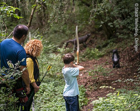 Drei Personen im Wald, ein Kind mit Bogen zielt auf ein Tierziel, zwei Erwachsene beobachten.