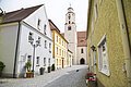 Ansicht einer alten Innenstadt mit bunten Häusern und einem Kirchturm im Hintergrund. Vor einigen Häusern stehen Pflanzgefäße und eine kleine Katze läuft auf der Straße.