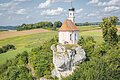 Kleine Kirche mit Zwiebelturm auf einem Felsen, umgeben von grünen Feldern und Bäumen.