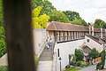 Eine Familie geht eine Treppe hinunter neben einer historischen Stadtmauer mit Ziegeldach, umgeben von grünen Bäumen.