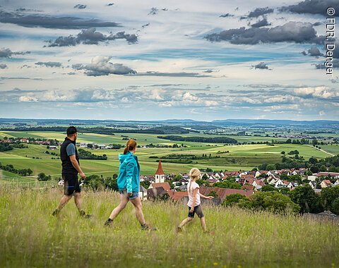 Geopark-Ries Juni 2022 Drei Personen wandern auf einem Hügel mit Blick auf eine ländliche Landschaft und ein Dorf im Hintergrund.
