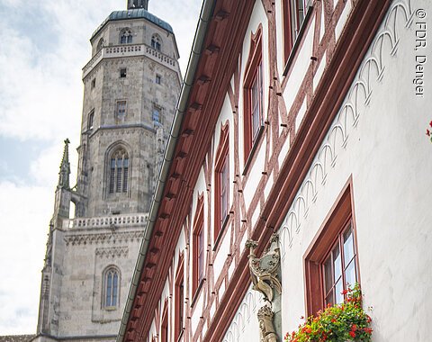 Fachwerkhaus mit roten Fensterrahmen und Blumenkästen, daneben ein Kirchturm.