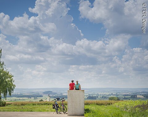 Zwei Personen sitzen auf einem Betonblock in einer Landschaft mit Fahrrädern daneben, Baum und Wolkenhimmel.