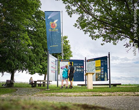 Zwei Personen betrachten Informationstafeln im Geopark Ries. Eine weitere Person sitzt auf einer Bank im Hintergrund.