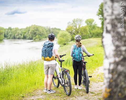 Zwei Personen mit Fahrrädern und Rucksäcken stehen auf einem Weg am Flussufer, umgeben von Bäumen.