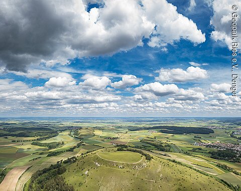 Weite Landschaft mit Feldern, Hügeln und einem Dorf unter einem Himmel mit vielen Wolken.