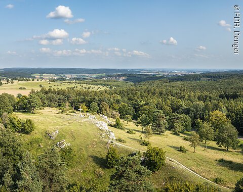 Drohnenfoto vom Bockberg bei Harburg mit grünen Hügeln und blauem Himmel. In der Mitte das Gipfelkreuz des Bockbergs