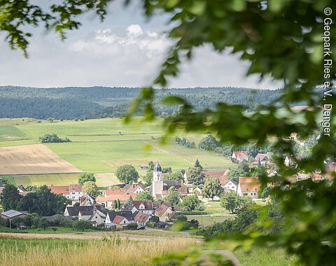 Landschaft mit Dorf, Feldern und Hügeln im Hintergrund, umrahmt von unscharfen Blättern im Vordergrund.