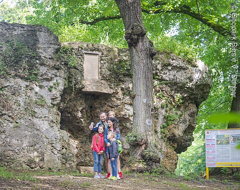 Eine Gruppe von Menschen steht vor einer großen Felsformation im Wald. Daneben befindet sich ein Informationsschild.