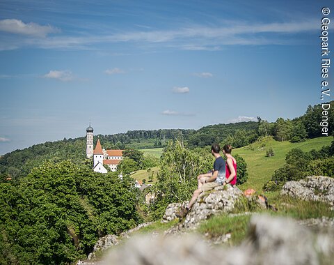 Zwei Personen sitzen auf einem Felsen und blicken auf eine Kirche in einer grünen Landschaft.