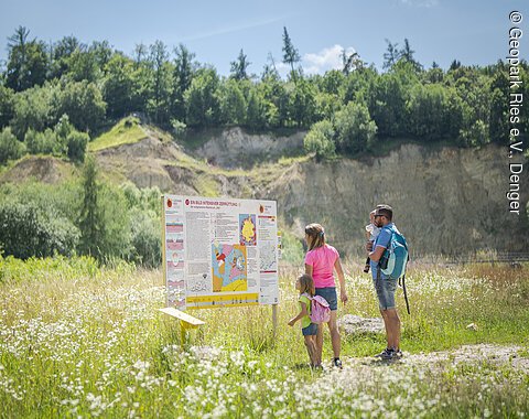 Eine Familie steht vor einem Informationsschild in einer grünen Landschaft mit Hügeln im Hintergrund.