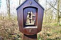 Kreuzwegstation bei der Brandkapelle in Monheim mit einer Holzstatue im Wald zeigt Jesus mit Kreuz. Oberhalb der Statue ein Kreuz, unterhalb die römische Nummer II. Hinter dem Wald bewölkter Himmel.