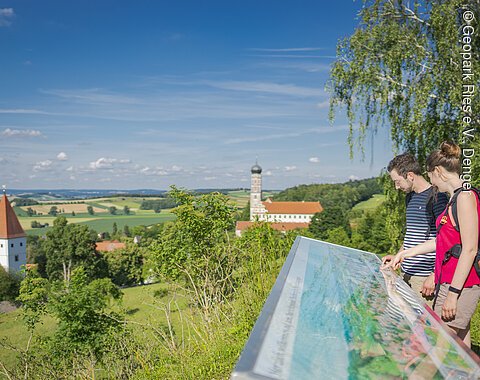 Zwei Personen betrachten eine Informationstafel mit Blick auf eine ländliche Landschaft mit Kirche und Kloster.