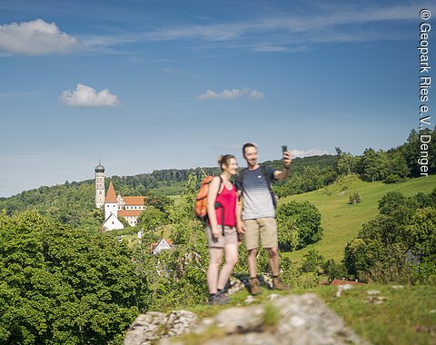 Zwei Personen machen ein Selfie auf einem Hügel mit einer Kirche und grüner Landschaft im Hintergrund.