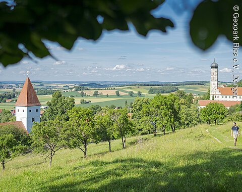 Landschaft mit Kirche, Feldern und zwei Personen, die auf einem Weg spazieren.