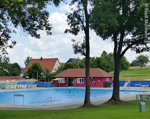 Ein Freibad mit Schwimmbecken, einer Rutsche und Sitzmöglichkeiten, umgeben von Bäumen und Wiesen. Im Hintergrund steht ein rotes Gebäude mit Fenstern, ein Kiosk.