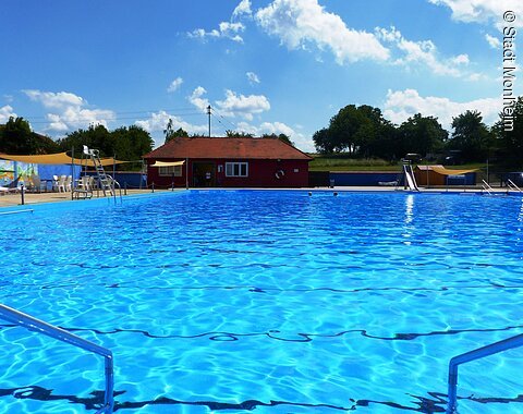 Das Freibad in Monheim: Schwimmbecken mit klarem Wasser, umgeben von Liegestühlen, einem roten Gebäude und Umkleidekabinen im Hintergrund.