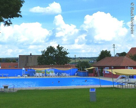 Freibad mit blauem Wasser, umgeben von Rasen, Bäumen und Gebäuden unter einem bewölkten Himmel. Vorderhalb vom Becken sind Sitzmöglichkeiten.