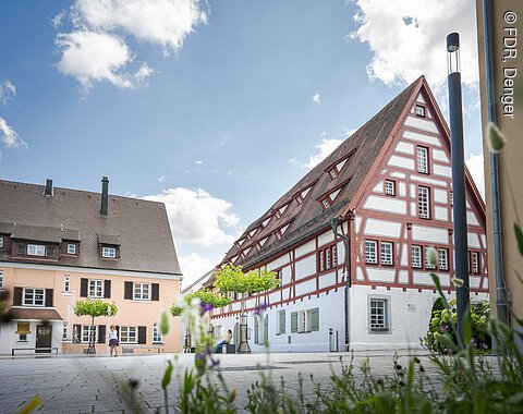 Fachwerkhaus mit rotem Giebel und weißen Wänden, daneben ein helles Gebäude, blauer Himmel im Hintergrund.