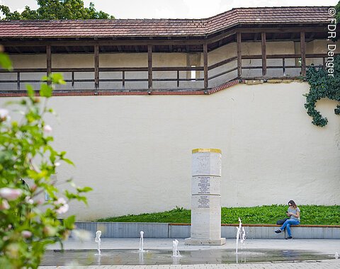 Eine Person sitzt auf einer Bank vor einer historischen Mauer mit Holzdach. Im Vordergrund sind Springbrunnen und eine Steinsäule.