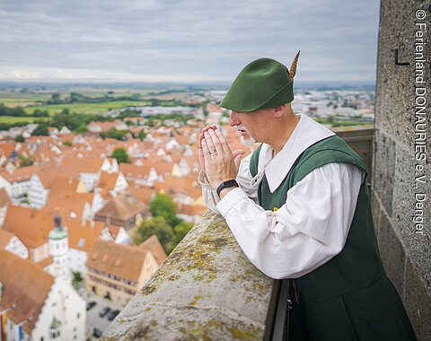 Person in historischer Kleidung steht auf einem Turm und blickt auf eine Stadt mit roten Dächern.