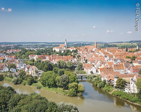Luftaufnahme einer Stadt mit roten Dächern, Fluss und Brücke, umgeben von grüner Landschaft.