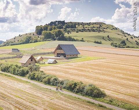 Landschaft mit Feldern, Hügeln und mehreren traditionellen Gebäuden. Zwei Radfahrer auf einem Weg im Vordergrund.
