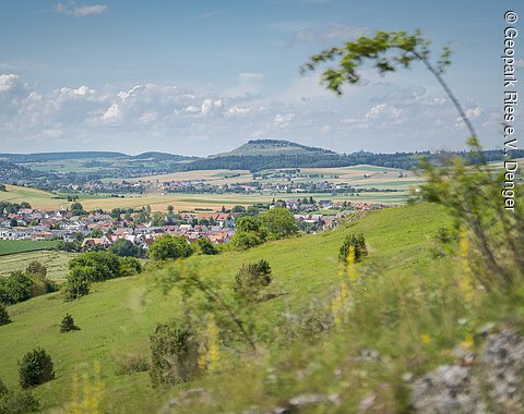 Landschaft mit grünen Hügeln, einem Dorf im Tal und einem bewaldeten Hügel im Hintergrund unter blauem Himmel.