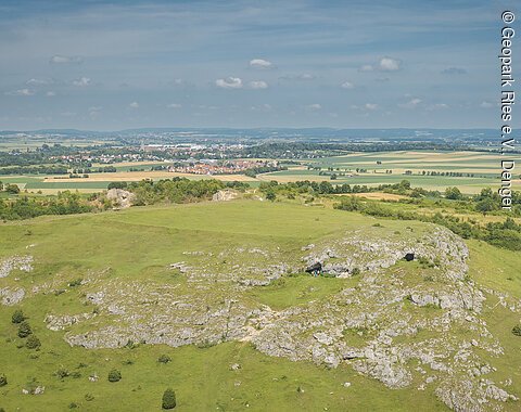 Hügelige Landschaft mit grünen Wiesen und Felsen im Vordergrund, Felder und ein Dorf im Hintergrund.