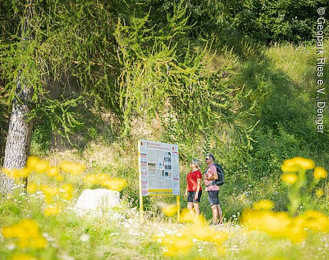 Zwei Personen stehen vor einem Informationsschild in einer grünen, blumigen Landschaft.