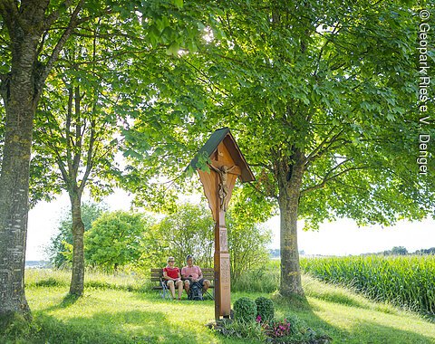 Zwei Personen sitzen auf einer Bank unter Bäumen neben einem hölzernen Wegkreuz.