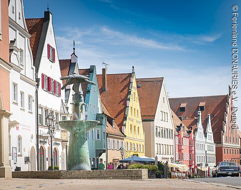 Straße mit bunten Giebelhäusern und einem Brunnen im Vordergrund. Klarer Himmel im Hintergrund.