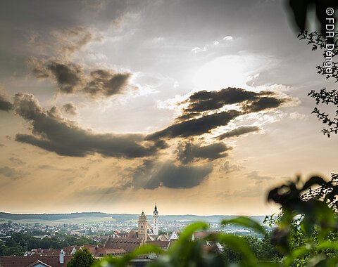 Stadtansicht mit Kirchtürmen im Vordergrund, dramatischer Himmel mit Sonnenstrahlen und Wolken.
