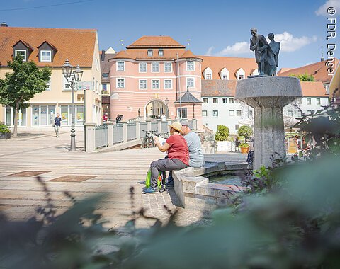 Personen sitzen auf einer Bank neben einem Brunnen auf einem Platz, im Hintergrund sind Gebäude mit roten Dächern.