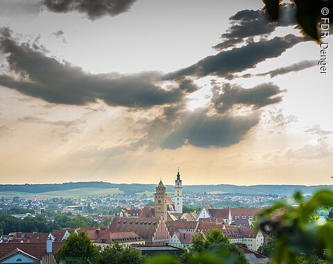 Stadtansicht Donauwörths mit Kirchtürmen der Liebfrauenkirche und des Klosters Heilig Kreuz im Vordergrund, bewölkter Himmel und Sonnenstrahlen im Hintergrund.