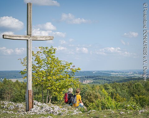 Bockberg Harburg Holzkreuz auf einem Hügel, zwei Personen mit Rucksäcken sitzen daneben, Blick auf bewaldete Landschaft.