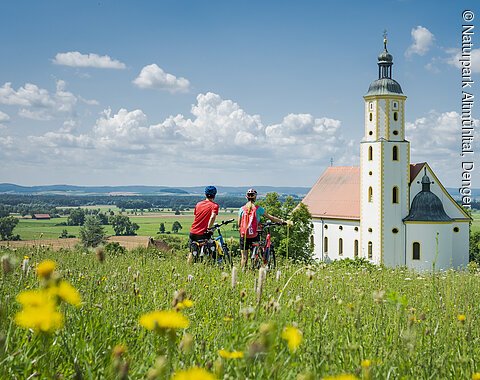 Zwei Radfahrer stehen auf einer Wiese vor einer Kirche in einer ländlichen Landschaft.