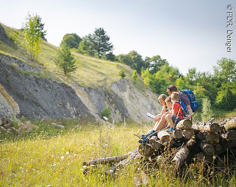 Drei Personen sitzen auf einem Stapel Baumstämme in einer hügeligen Landschaft und lesen in einem Buch.