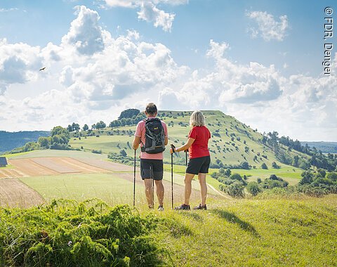 Zwei Personen mit Wanderstöcken stehen auf einem Hügel und blicken auf eine ländliche Landschaft.
