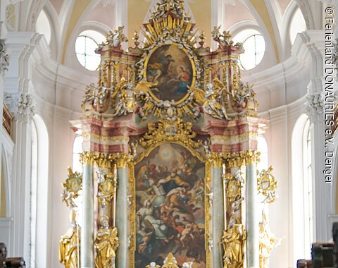 Zwei Personen mit Rucksäcken stehen in einer Kirche vor einem reich verzierten Altar.