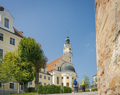 Zwei Personen mit Rucksäcken gehen auf eine gelbe Kirche mit Turm zu, umgeben von Bäumen.